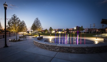 Photograph of an urban park at dusk featuring a circular splash pad with brightly colored, illuminated water jets surrounded by a curved brick seating wall and wide concrete walkways.