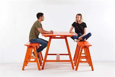 tudio photograph of a bright orange, modern picnic table with two attached backless benches. Two people are sitting on the benches facing each other against a solid white background.